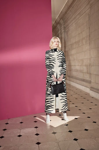 A fair-skinned woman with blond hair stands in a zebra-patterned coat, holding a black purse in an indoor setting against a pink wall.