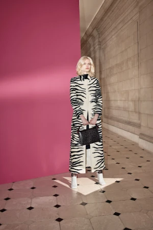 A fair-skinned woman with blond hair stands in a zebra-patterned coat, holding a black purse in an indoor setting against a pink wall.