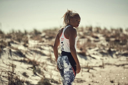Adult female in fitness wear stands on a sandy beach, looking back over her shoulder at the camera, with dunes in the background.