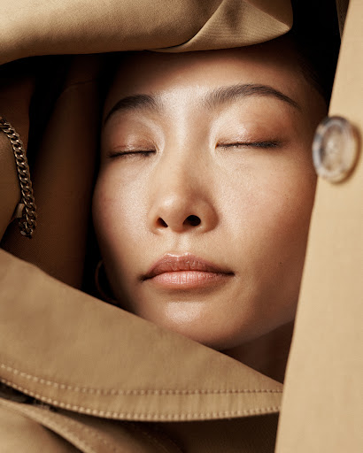Close-up portrait of a serene Asian adult female with closed eyes, framed by a tan trench coat, accessorized with a gold bracelet.