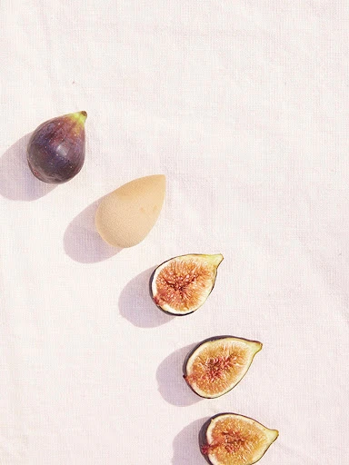 Overhead shot of a halved figs and a makeup sponge on a white textured fabric background. Food photography, beauty product image.