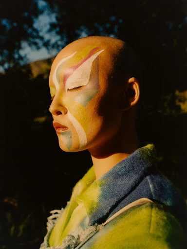 Portrait of a woman with bald head and artistic makeup, posing in a green coat against a nature background, eyes closed, illuminated by sunlight.