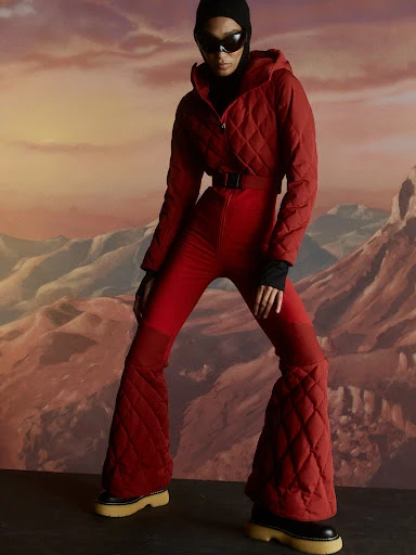 A person in a red quilted ski suit, black balaclava, and black sunglasses poses in front of a landscape with mountains.