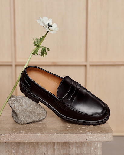 Black leather penny loafer shoe is showcased on a stone surface with a white flower, set against a beige paneled backdrop.