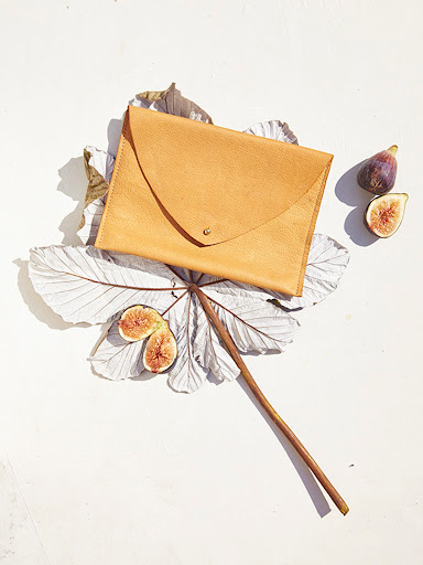 Tan leather clutch resting on dried leaves with sliced figs against a light textured surface