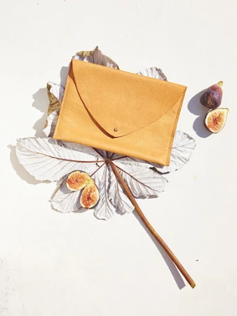 Tan leather clutch resting on dried leaves with sliced figs against a light textured surface