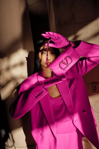 An Asian model poses indoors with a Valentino purse wearing a pink suit, pink shirt, and pink gloves in a fashion editorial.