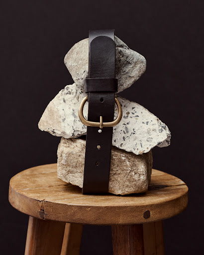 Dark brown leather belt with a gold buckle wrapped around stacked grey rocks on top of a round wooden stool against a dark background.