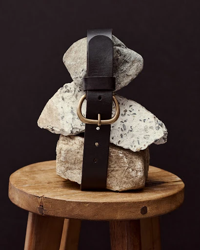 Dark brown leather belt with a gold buckle wrapped around stacked grey rocks on top of a round wooden stool against a dark background.