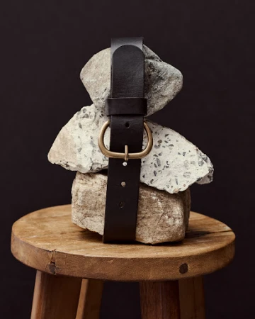Dark brown leather belt with a gold buckle wrapped around stacked grey rocks on top of a round wooden stool against a dark background.