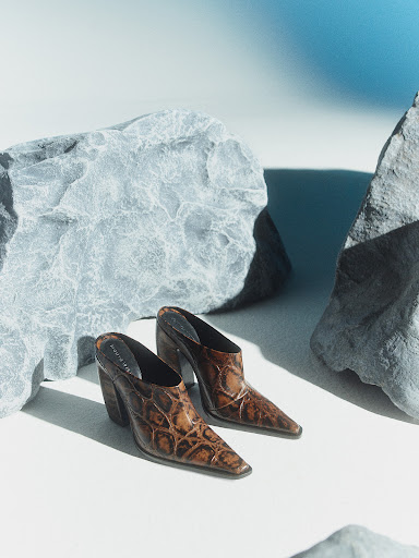 Two brown, crocodile-patterned heeled mules are showcased with a light blue and white background and surrounded by rough, gray rocks.