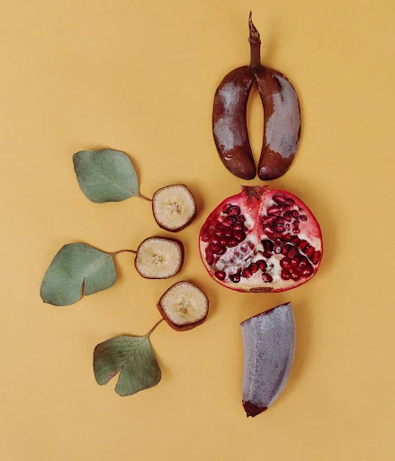 Still life studio shot featuring a halved pomegranate, sliced kiwi, overripe banana, and leaves against a pastel yellow backdrop.
