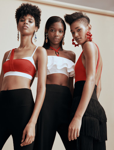 Three women pose in a studio wearing red and white crop tops with black pants and statement earrings, against a neutral backdrop.