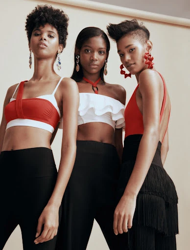 Three women pose in a studio wearing red and white crop tops with black pants and statement earrings, against a neutral backdrop.