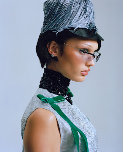 Model wearing a feather hat and embellished high neck in a studio portrait against a white background, fashion editorial shot.