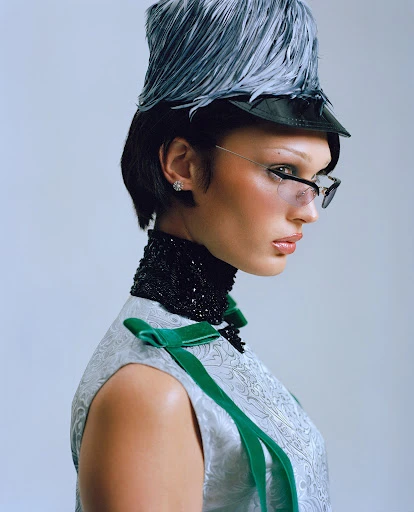 Model wearing a feather hat and embellished high neck in a studio portrait against a white background, fashion editorial shot.