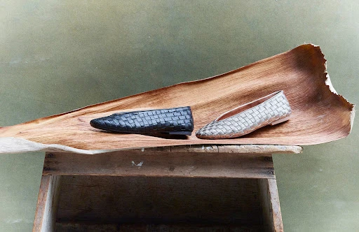 Still life of woven leather flats, one black, one silver, displayed on a large dried leaf atop a rustic wooden crate indoors.