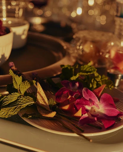 Arrangement of purple orchids, mint sprigs, and sliced citrus on a plate with a blurred background showcasing glasses and dishware.