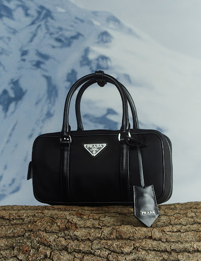 Prada black handbag on log against blurred snowy mountain backdrop. Luxury fashion accessory with logo detail and leather tag.