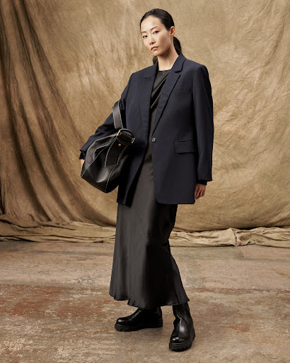 Adult woman in an oversized navy blazer, black dress, black boots, and carrying a black shoulder bag, posing against a mottled brown backdrop.