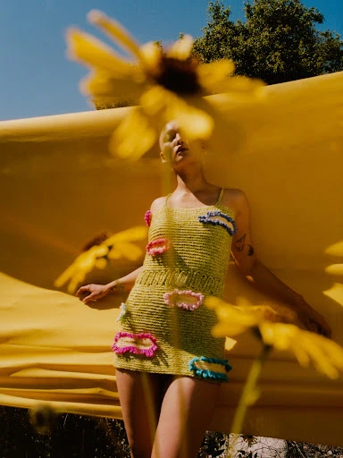 Woman in a yellow crocheted dress poses outdoors with bright yellow flowers and a matching fabric backdrop under a blue sky.