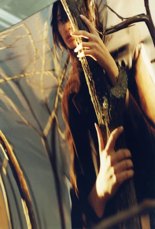 Woman posing by a tree trunk with dappled light. The image features soft lighting and a serene atmosphere with natural elements.