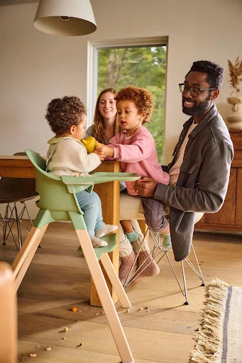Indoor family scene featuring a Black father, Caucasian mother, and two children, one in a high chair, sharing food at a dining table.