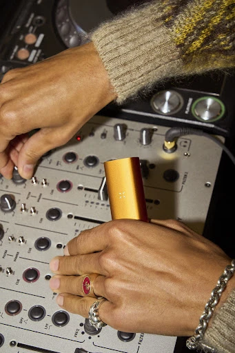 Close-up of tan-skinned hands adjusting knobs on a DJ mixer, wearing rings and a bracelet. A gold vaporizer stands upright.