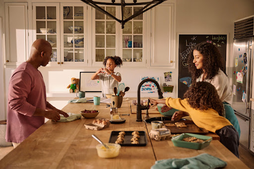 An African-American family of four baking cookies together in a sunlit kitchen with white cabinets, a large wooden table, and chalkboard art.