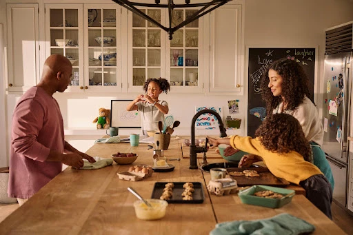An African-American family of four baking cookies together in a sunlit kitchen with white cabinets, a large wooden table, and chalkboard art.