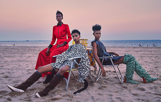 Three fashion models pose in colorful outfits on a sandy beach at dusk, seated on beach chairs with ocean backdrop.