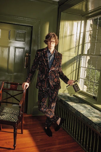 A young adult male poses in a patterned suit with a vintage radio indoors by a window. The scene is lit by natural light.