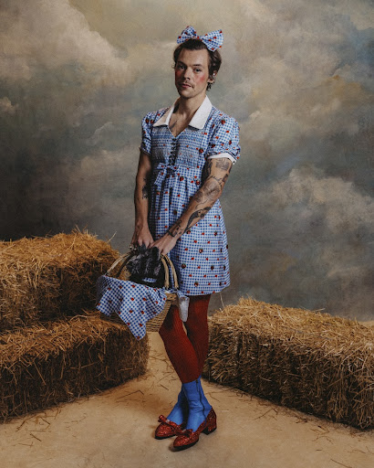 Portrait of a male in a blue and white gingham dress with a matching bow, holding a basket, with hay bales in the background.