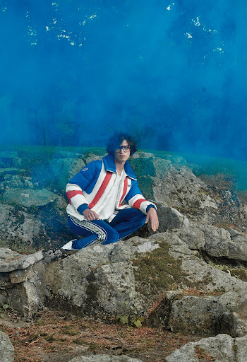 Man in a blue and white jacket and blue pants poses on rocks with a blue smokey background in a fashion editorial shot.