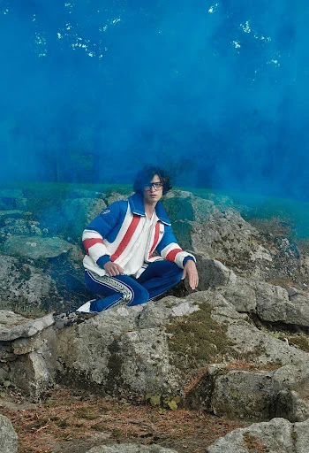 Man in a blue and white jacket and blue pants poses on rocks with a blue smokey background in a fashion editorial shot.