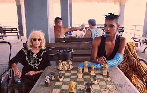 Two women at a checkerboard table near a beach. One woman is elderly, wearing sunglasses. The other is a younger fashion model.