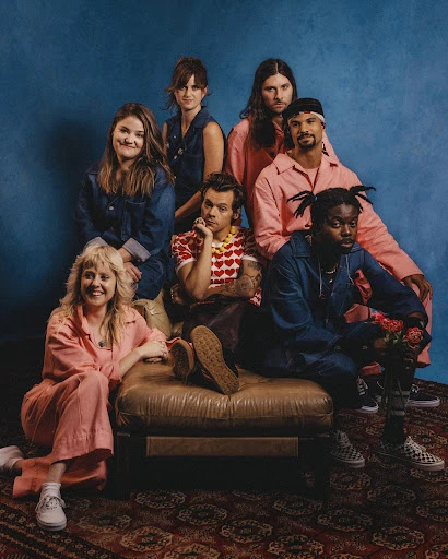 Group portrait of seven adults wearing casual clothing, posing indoors against a blue wall with a printed rug and a brown leather sofa.