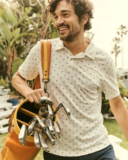 Smiling man carrying a golf bag with clubs. He wears a patterned polo shirt and navy shorts in an outdoor setting with palm trees.