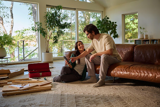 A couple assembles furniture in a bright, modern living room with a large window overlooking a serene lake landscape, promoting home improvement.