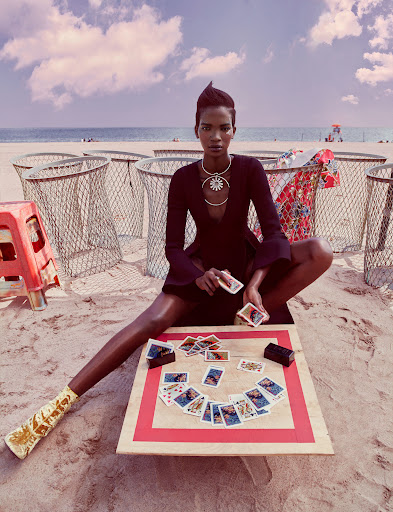 Black woman seated on a sandy beach holding playing cards before a small table with spread cards