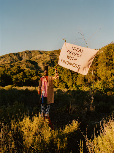 Adult stands in a field holding a flag that reads TREAT PEOPLE WITH KINDNESS, wearing a coat, and the scene is set on a rural hillside.