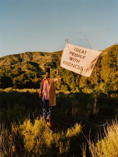 Adult stands in a field holding a flag that reads TREAT PEOPLE WITH KINDNESS, wearing a coat, and the scene is set on a rural hillside.