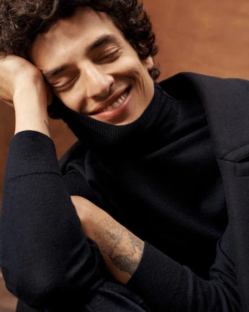 Close-up portrait of a smiling man with curly dark hair wearing a black turtleneck and resting his head on his hand.