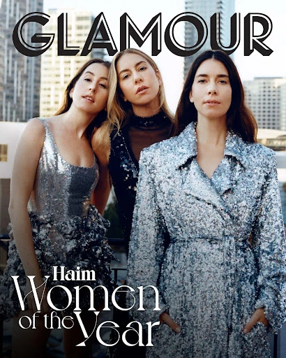 Glamour magazine cover featuring the Haim sisters in sparkly dresses and coats, standing against a city skyline backdrop.