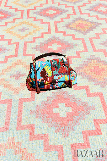 Stylized fashion handbag with colorful textile design sits on a patterned pink and orange textile background, featuring a Bazaar logo.