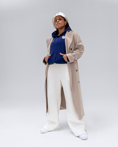 Full shot of a woman in a navy hoodie, white pants, sneakers, and a trench coat posing against a neutral background in a studio.