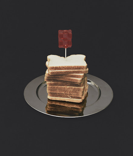 Stack of bread slices on a silver plate topped with a gummy candy on a stick, set against a dark grey background in studio lighting.