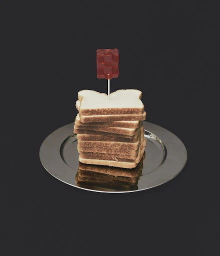 Stack of bread slices on a silver plate topped with a gummy candy on a stick, set against a dark grey background in studio lighting.