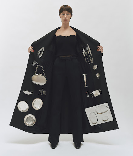 Stylish woman in black coat presents silver tableware and plates. Studio fashion portrait on white background. Minimalist, creative.