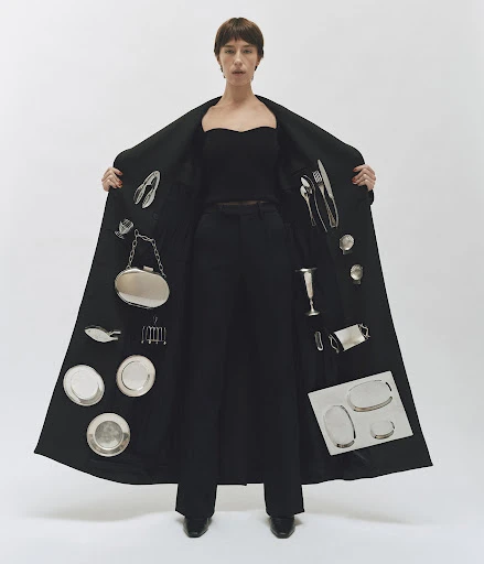Stylish woman in black coat presents silver tableware and plates. Studio fashion portrait on white background. Minimalist, creative.